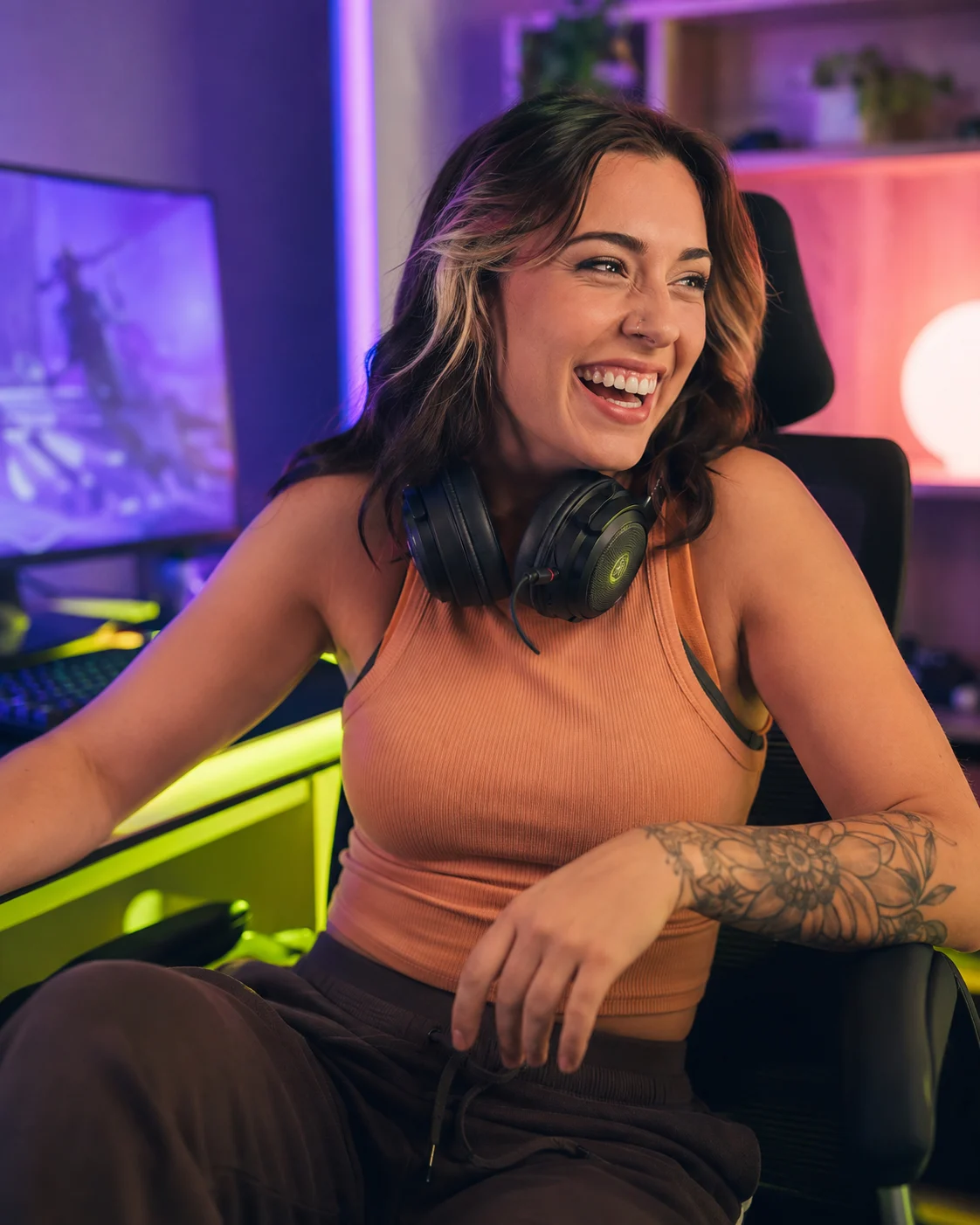 An adult gamer with a headset around her neck laughing at her desk in violet and lime light